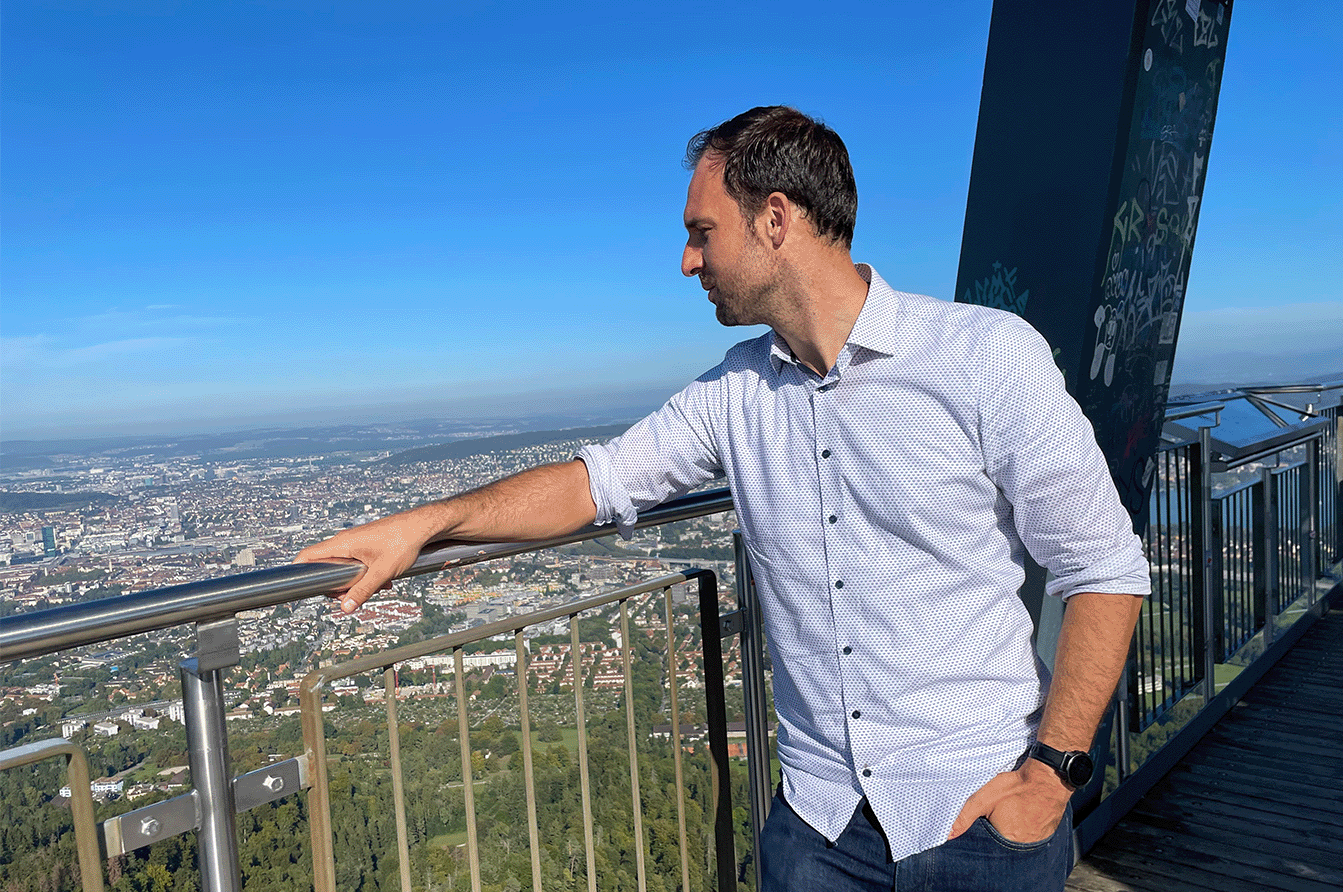 Ein Mann steht auf einer Aussichtsplattform. Seine Hand liegt auf dem Geländer und er blickt nach rechts auf die Aussicht auf die Stadt Zürich.
