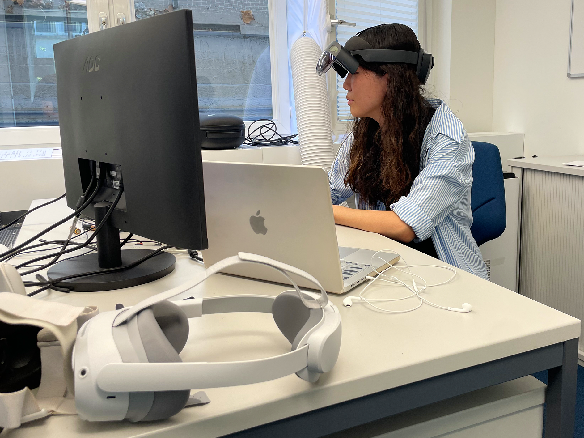 Portia is sitting at her desk and wears AR glasses. In the foreground of the picture are a pair of VR glasses.