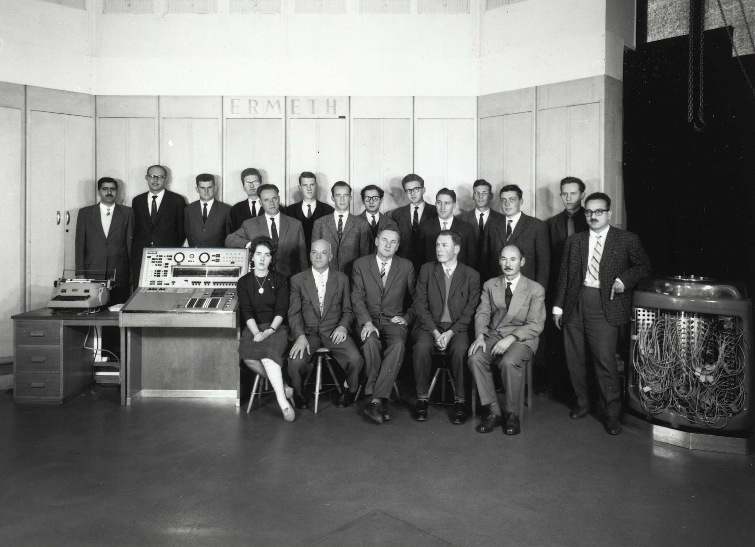 Enlarged view: Black and white photo of a group of people posing in front of tall wooden cabinets that read "ERMETH". On the left is an old console, on the right a large magnetic drum with many cables.